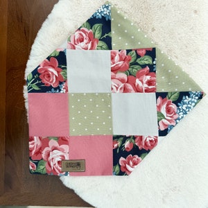 May include: A patchwork bandana featuring a floral design in shades of pink, green, white and blue. The bandana is made from cotton fabric and has a square shape. The bandana has a brown leather tag with the text "Cheeky Beanie" on it.