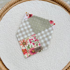 May include: A patchwork bandana featuring a floral print, gingham pattern, and polka dots in shades of pink, green, and white. The bandana has a brown leather tag with the text "Cheeky & Brave" on it.