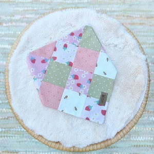 May include: A pink, green and white patchwork bandana featuring strawberries, bees and flowers. The bandana has a brown label with the text "BEE KIND" on it.