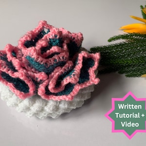 Könnte beinhalten: Eine gehäkelte Blume aus rosa und türkisfarbenem Garn, geschichtet auf einer weißen gehäkelten Basis. Das Bild enthält den Text "Schriftliches Tutorial + Video".