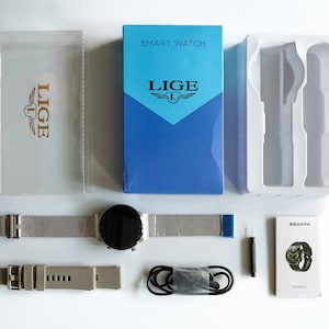 Op de afbeelding: Een zilveren smartwatch met een zwart display en een metalen band. Het horloge wordt geleverd in een blauwe doos met het woord "LIGE" erop gedrukt. De doos bevat ook een witte plastic behuizing, een zwarte oplaadkabel, een kleine schroevendraaier en een handleiding.