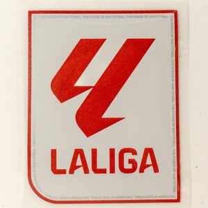 Puede incluir: Una pegatina blanca con un logotipo rojo de una L estilizada y la palabra "LALIGA" en rojo. El texto "THE POWER OF OUR FUTBOL" se repite alrededor del borde de la pegatina.