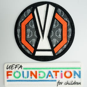 May include: Two embroidered patches, one round and one rectangular. The round patch is black with a white and orange design of a trophy. The rectangular patch is white with the text "UEFA FOUNDATION for children" in colourful letters.
