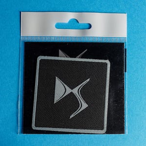 Könnte beinhalten: Ein schwarzer und silberner gestickter Aufnäher mit dem DS Automobiles Logo. Der Aufnäher befindet sich in einer durchsichtigen Plastiktüte mit einem Aufhängeloch.