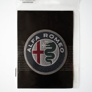 May include: A close-up of a gray and black embroidered patch with the Alfa Romeo logo. The logo features a red cross and a green snake.