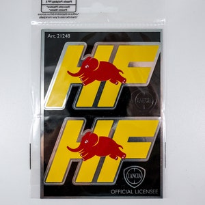 Può includere: Due adesivi gialli e argentati con un logo di elefante rosso e il testo "HF" e "LANCIA" su sfondo nero. Gli adesivi sono confezionati in un sacchetto di plastica trasparente con il testo "Art. 2 1248" e "OFFICIAL LICENSEE".