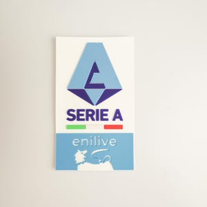 Può includere: Un logo blu e viola con il testo "SERIE A" e una bandiera italiana sotto. Il logo è una lettera stilizzata "A" con una lettera "C" più piccola all'interno. Il testo "enilive" è sotto la bandiera.