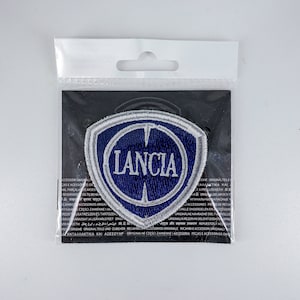 Può includere: Una toppa con il logo Lancia blu e bianco in un sacchetto di plastica trasparente. La toppa è a forma di scudo con la scritta "LANCIA" in lettere bianche. La toppa è su sfondo nero.
