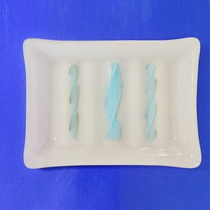 May include: A rectangular white soap dish with three vertical ridges. The dish features three light blue, twisted glass accents. The soap dish is made of glass and has a glossy finish.