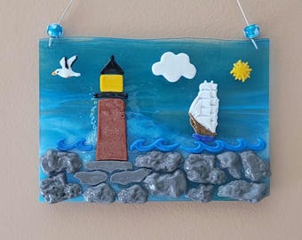 May include: A rectangular glass art piece depicting a coastal scene. It features a lighthouse with a yellow light, a sailboat, a seagull, a white cloud, and a yellow sun. The scene includes blue waves and gray rocks.