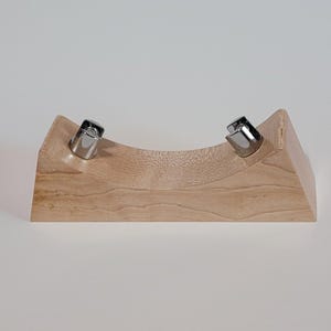 May include: A wooden display stand with a curved center and two silver metal clips. The stand is made of light-colored wood and has a simple, modern design. The clips are positioned to hold an item.