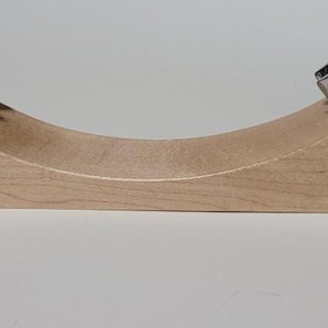 May include: A wooden wine bottle holder with a curved design. The holder is made of light-colored wood and features two metal fixtures to secure the bottle. The holder is designed to cradle a wine bottle.