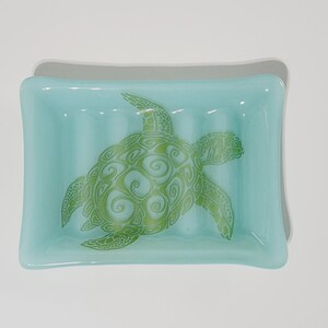 May include: A rectangular, light blue soap dish with a green sea turtle design. The turtle is detailed with swirling patterns. The dish has a slightly curved shape and is made of glass.