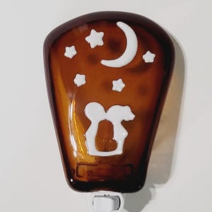 May include: Brown night light with a white moon, stars, and a couple silhouette design. The night light has a rectangular shape with a rounded top and a white plug-in base. A decorative accent for any room.