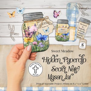 Puede incluir: Un tarro de albañilería de papel con un diseño floral, mariposas y el texto "Sweet Meadow Hidden Paperclip Secret Note Mason Jar". El tarro está sostenido por una mano. Incluye clips y otros elementos decorativos.