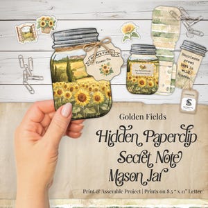 May include: A DIY paper craft project featuring a mason jar cutout with a sunflower field illustration, a tag labeled Golden Fields Flower Co., and a smaller jar cutout with the text Happiness grows best in wild spaces, plus metal paperclips.