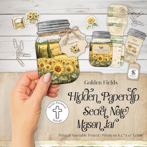 May include: A paper craft project featuring a mason jar illustration with a sunflower field scene, a tag labeled Fields of Grace Flower Co., and a note with the text His goodness blooms in every season, accompanied by metal paperclips on a wood background.
