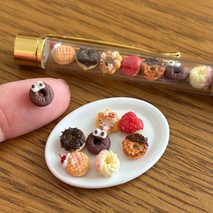 以下が含まれることがあります： 透明なチューブと白いお皿に並んだミニチュアのドーナツ。チョコレート、イチゴ、グレーズなど、様々な色とスタイルがあります。指の上にもドーナツが一つ。ドールハウスやクラフトプロジェクトに最適です。
