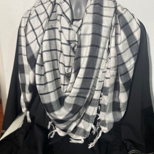 May include: A black and white checkered scarf with fringed edges is draped over a black garment. The scarf is made of a lightweight fabric and features a classic plaid pattern. The scarf is displayed on a mannequin.
