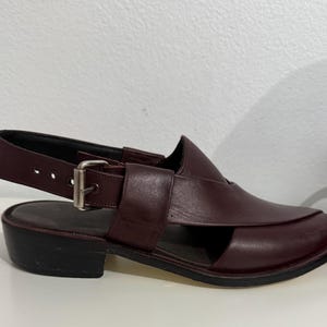 May include: Burgundy leather sandal with a square toe and an ankle strap with a buckle. The shoe has a low, block heel and a cut-out design. The sandal is a slip-on style.