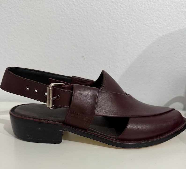 May include: Burgundy leather sandal with a square toe and an ankle strap with a buckle. The shoe has a low, block heel and a cut-out design. The sandal is a slip-on style.