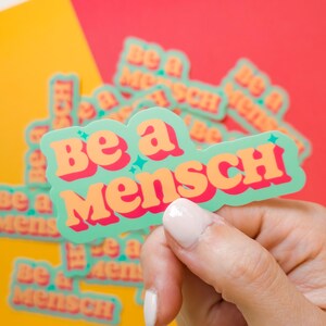K&ouml;nnte beinhalten: Ein hellgr&uuml;ner Aufkleber mit dem Schriftzug "Be a Mensch" in orangefarbenen Buchstaben mit roter Umrandung. Der Text ist mit t&uuml;rkisfarbenen Sternen verziert. Weitere &auml;hnliche Aufkleber befinden sich im Hintergrund.