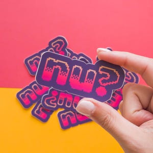 May include: A collection of stickers with the word "nü?" in a pixelated font, held by a hand. The stickers have a purple outline with pink and orange pixelated text. The background is split between red and yellow.