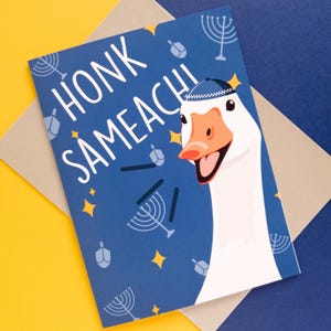 May include: A greeting card with a blue background featuring the text "HONK SAMEACHI" and a cartoon goose wearing a yarmulke. The card includes illustrations of menorahs, dreidels, and stars. The card is on a yellow and blue surface.