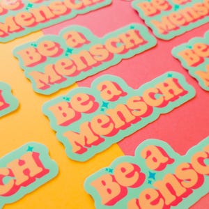 K&ouml;nnte beinhalten: Mehrere Aufkleber mit dem Schriftzug "Be a Mensch" in Orange, umrandet mit Rot, auf hellgr&uuml;nem Hintergrund. Die Aufkleber sind auf einem gelben und rosa Hintergrund angeordnet.