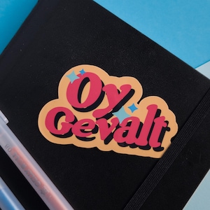 May include: A black notebook with a sticker that says "Oy Gevalt" in pink letters with a yellow outline and blue star accents. Two pens are visible in the lower left corner.