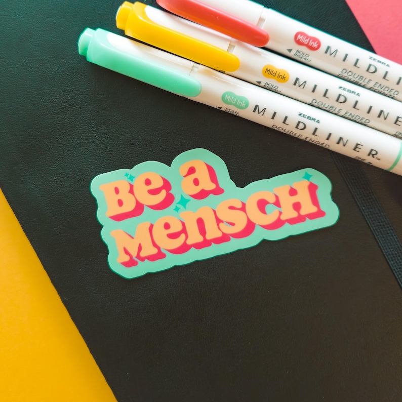 K&ouml;nnte beinhalten: Ein schwarzes Notizbuch mit einem Aufkleber mit der Aufschrift "Be a Mensch" in Orange und Rosa. Drei Zebra Mildliner Textmarker mit doppelter Spitze in Orange, Gelb und Mintgr&uuml;n liegen auf dem Notizbuch.