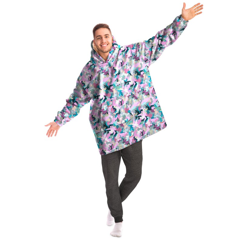 May include: A person wearing a large, oversized hoodie with a pink, blue, and green camouflage pattern. The hoodie has a drawstring hood and kangaroo pocket. The person is wearing grey sweatpants and white socks.
