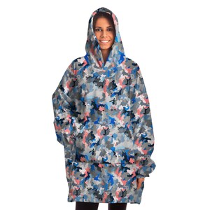 Puede incluir: Una manta con capucha con un patr&oacute;n de camuflaje azul, rosa y gris. La manta tiene un bolsillo grande en la parte delantera y una capucha con cord&oacute;n.
