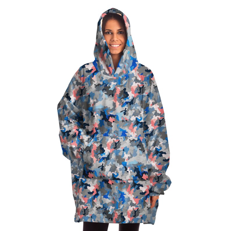 Puede incluir: Una manta con capucha con un patr&oacute;n de camuflaje azul, rosa y gris. La manta tiene un bolsillo grande en la parte delantera y una capucha con cord&oacute;n.