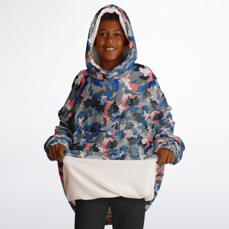 Puede incluir: Una manta port&aacute;til con capucha de color blanco con forro de felpa y estampado de camuflaje azul, gris y rosa. La manta tiene una capucha y un bolsillo grande en la parte delantera.