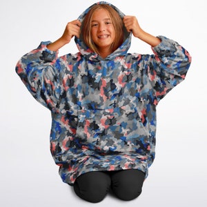 Puede incluir: Una manta con capucha de camuflaje azul, rosa y gris con un gran bolsillo en la parte delantera. La manta est&aacute; hecha de un tejido suave y afelpado y es perfecta para acurrucarse en el sof&aacute;.