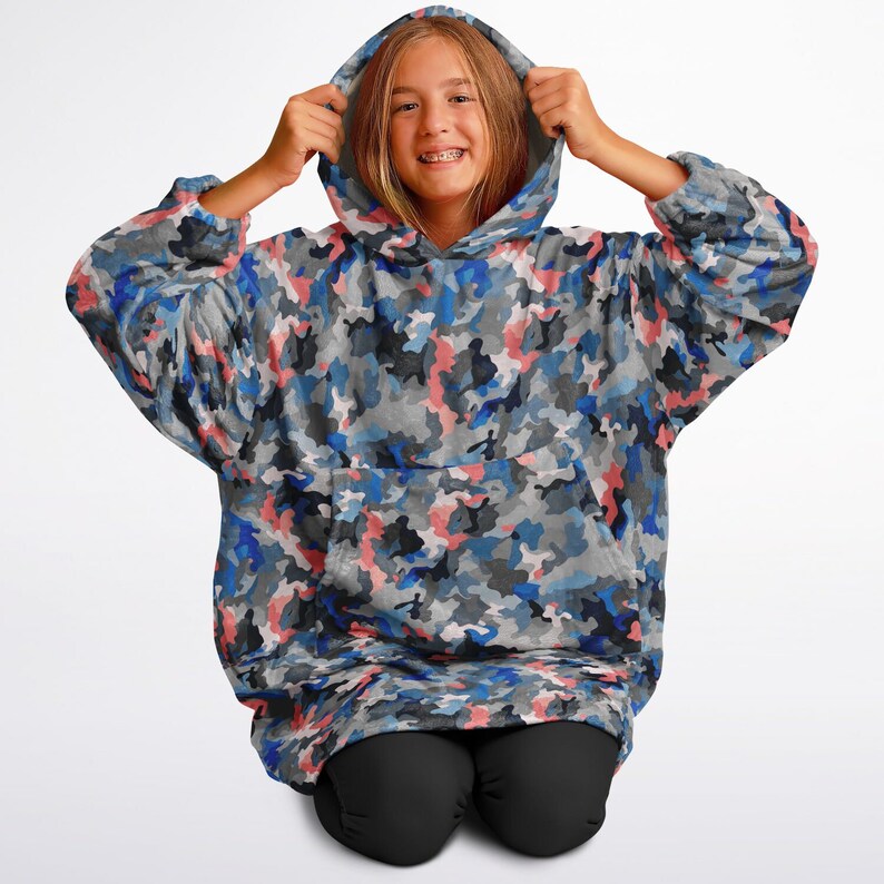 Puede incluir: Una manta con capucha de camuflaje azul, rosa y gris con un gran bolsillo en la parte delantera. La manta est&aacute; hecha de un tejido suave y afelpado y es perfecta para acurrucarse en el sof&aacute;.