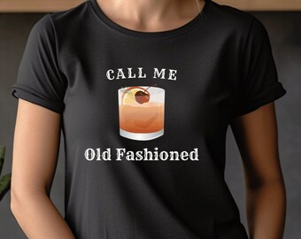 May include: Black t-shirt with the text "CALL ME Old Fashioned" and a graphic of an Old Fashioned cocktail. The shirt is worn by a person. The background includes a wooden shelf and plants.