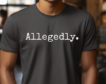 May include: Dark gray t-shirt with the word "Allegedly." printed in white, displayed in a casual font. The shirt is short-sleeved and appears to be made of a soft material. The person is wearing the shirt with their hands in their pockets.