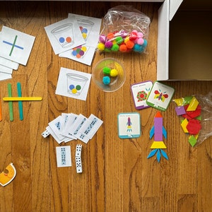 May include: A set of educational toys for children, including colorful pom poms, wooden sticks, and cards with different shapes and patterns. There are also cards with numbers and a lion puzzle.