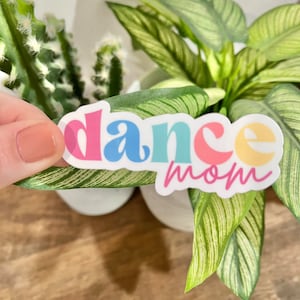 May include: A colorful sticker with the text "dance mom" in a rainbow font. The sticker is on a green plant.