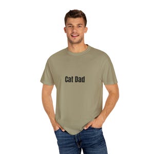 Puede incluir: Una camiseta color caqui con el texto "Cat Dad" impreso en la parte delantera.