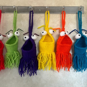 May include: Six crocheted hanging pockets with googly eyes and fringe. The pockets are in the colors red, green, purple, yellow, orange, and blue.