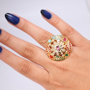 May include: A gold-toned ring with a floral design, encrusted with various colored gemstones. The ring is set on a hand with dark blue nail polish. The ring's design includes red, green, blue, and pink stones.