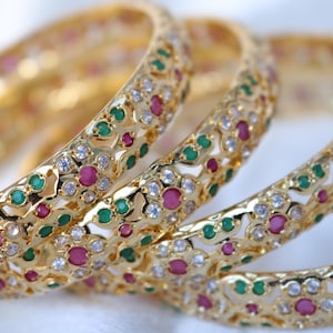 May include: Close-up of several gold bangles. Each bangle is adorned with a repeating pattern of small, round, red and green gemstones, interspersed with clear, sparkling stones. The bangles are arranged in a slightly overlapping manner, showcasing their intricate design.