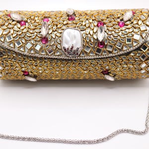 May include: A gold-coloured clutch bag with a silver chain strap. The bag is decorated with small mirrors, pink and clear gemstones. The clasp features a large, pearlescent stone with a dark red centre.