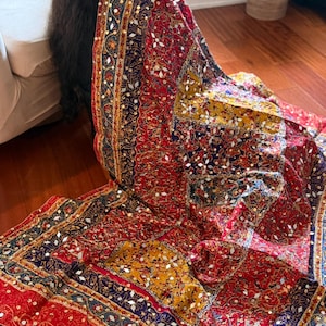 May include: A rectangular textile wrap featuring a vibrant patchwork design in shades of red, navy blue, and mustard yellow. The fabric is heavily embellished with intricate gold-toned embroidery and small, reflective mirror-like circular accents.