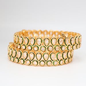 May include: Two gold bangles with a repeating pattern of teardrop-shaped clear stones and green accents. The bangles are stacked on top of each other, showcasing the intricate design and craftsmanship. The jewelry is set against a plain white background.