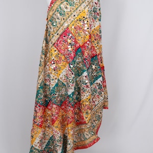 May include: A colorful, patterned scarf with a patchwork design. The scarf features a vibrant mix of red, yellow, green, and white squares, adorned with floral patterns and small reflective embellishments. The edges are trimmed with a decorative border.