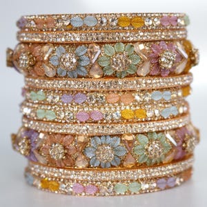 May include: Stack of ornate gold bangles with floral and gemstone embellishments. The bangles feature a variety of pastel colors, including pink, blue, yellow, and green, and are encrusted with sparkling rhinestones. The design is intricate and detailed.
